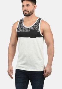 Hvid tanktop med sort mønstret brystpanel, lomme detalje og rund halsudskæring. Serveret med mørke denim jeans.