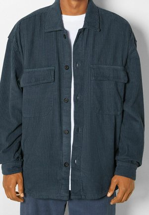 Homme portant une veste boutonnée en velours côtelé bleu foncé avec deux poches poitrine sur une chemise blanche.