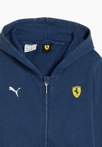 Felpa zip in navy blue realizzata in un tessuto morbido, con un logo bianco Puma sul petto e uno stemma giallo Ferrari sul cappuccio.
