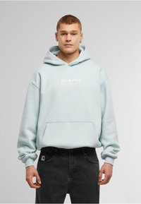 Felpa azzurra con cappuccio e tasca frontale, con un logo bianco con la scritta "KANI The Originator" sul petto. Jeans neri con patch del logo.