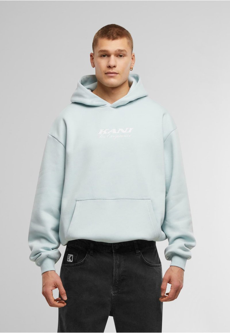 Felpa azzurra con cappuccio e tasca frontale, con un logo bianco con la scritta "KANI The Originator" sul petto. Jeans neri con patch del logo.