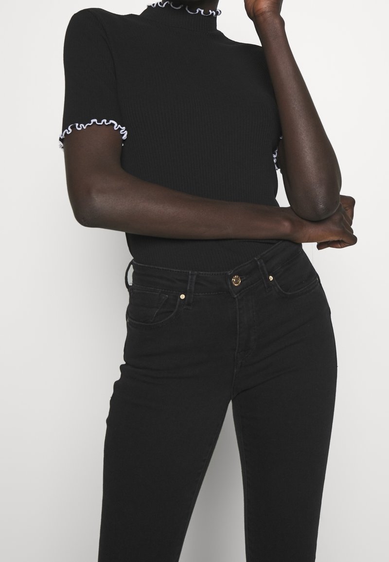 Persona que lleva una camiseta negra de manga corta con ribetes blancos y jeans negros de tiro alto con detalles de botones dorados.