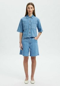 Camicia di jeans azzurro chiaro a maniche corte, con chiusura frontale a bottoni e due taschini sul petto; abbinata a pantaloni corti in jeans dello stesso colore e scarpe beige.