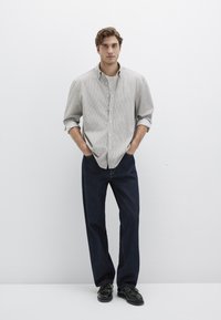 Hell gestreiftes Hemd über einem cremefarbenen Oberteil, kombiniert mit dunkelblauen, gerade geschnittenen Jeans und schwarzen Schuhen, die Hände in den Taschen, stehend vor einem weißen Hintergrund.