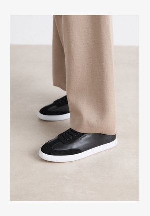 Zapatillas de cuero negro con acentos de ante, con una suela de goma blanca y cordones negros. Combinadas con pantalones de pierna ancha beige.