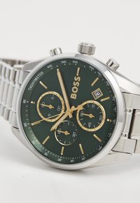 Silver stainless steel chronograph watch with a green dial, gold accents, and three sub-dials. Features a date window and tachymeter markings.