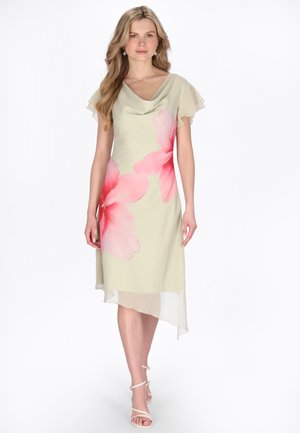 Kobieta w jasnobeżowej sukience z dużymi różowymi kwiatowymi nadrukami, z falbanowymi rękawami, asymetrycznym dołem oraz beżowych sandałach na obcasie, stojąca na białym tle.