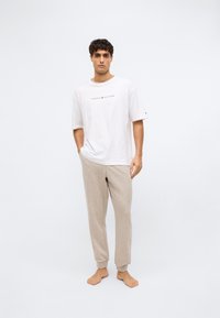 Witte T-shirt met het "TOMMY HILFIGER" logo in het midden, gecombineerd met lichtbeige sweatpants met elastische boorden. Model staat blootsvoets.