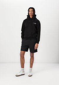 Svart hettegenser med frontlomme og snor, kombinert med svarte shorts. Modellen har på seg hvite joggesko med grå detaljer og sokker.
