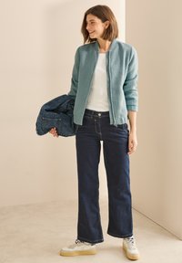 Bomber jacket azzurro chiaro, texture, indossato sopra un top bianco, abbinato a jeans larghi scuri e sneakers bianche con suola gialla.