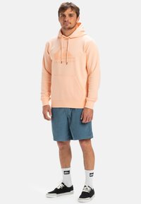 Light peach hoodie with drawstrings and front pocket, paired with teal shorts. Black shoes and white socks, featuring logo accents.