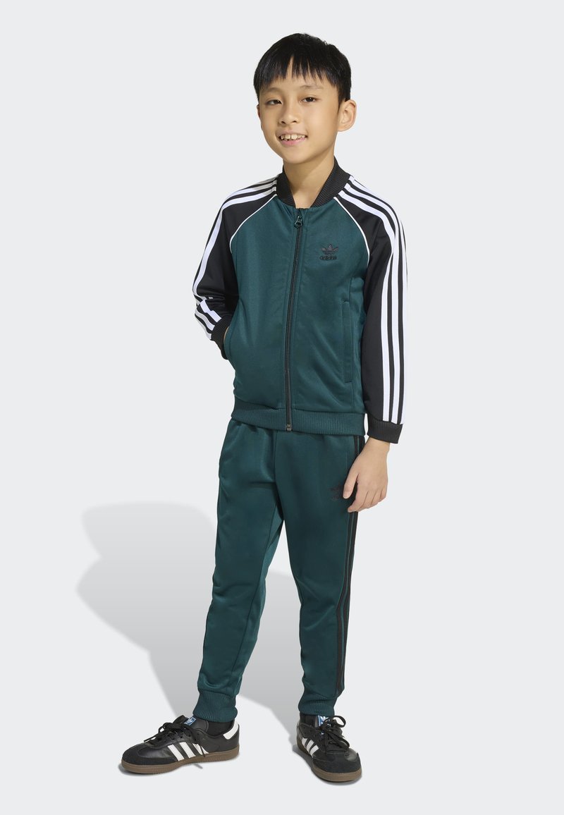 Grön träningsoverall med en zip-up jacka med svarta ärmar och tre vita ränder, i kombination med matchande byxor och svarta sneakers.