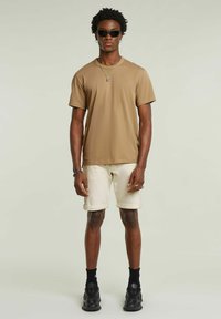 T-shirt in cotone color sabbia con collo girocollo, abbinata a pantaloni corti beige chiaro. Il modello indossa occhiali da sole neri e sneaker nere, con hardware minimale.