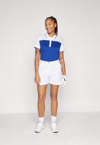 Camisa polo de golfe com um corpo azul e mangas brancas, combinada com calções brancos. Inclui um colarinho com botões e detalhes em malha.