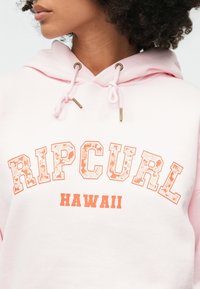 Sweat à capuche rose clair avec "RIPCURL HAWAII" en lettres orange audacieuses et à motif. Les cordons de serrage avec des finitions dorées ajoutent une touche de détail au design.