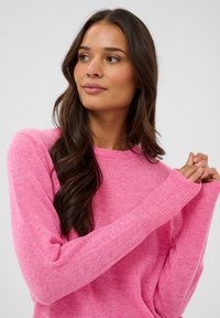 Pull en tricot rose avec des poignets côtelés. Il comporte un col rond et une texture douce. Le modèle pose avec les mains jointes près de la poitrine.