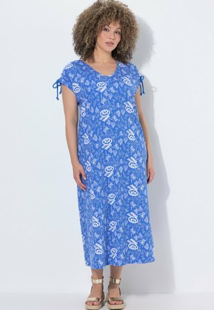 Frau mit lockigem Haar, die ein blau gemustertes Midikleid mit kurzen Ärmeln und gebundenen Details trägt, kombiniert mit goldenen Sandalen mit Knöchelriemen.