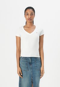 T-shirt blanc en coton à manches courtes avec un design en V, arborant un petit logo brodé sur la poitrine. Porté avec une jupe en denim bleu.