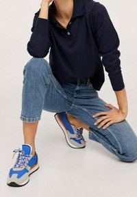 Sneakers sportive con una combinazione di colori grigia, blu e arancione, con una tomaia in tessuto testurizzato e suola in gomma, abbinate a jeans con risvolto e una maglietta blu navy.