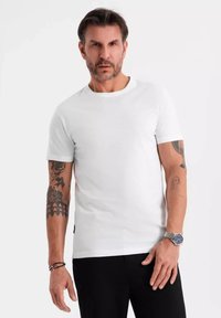 Hvid bomulds t-shirt med rund hals og korte ærmer, båret af en mand med synlige tatoveringer, parret med sorte bukser og et sølvur.