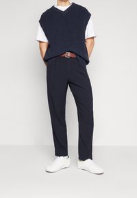 Pull sans manches en tricot marine porté sur un T-shirt blanc, associé à un pantalon sombre et des baskets blanches. Une ceinture brune accentue la tenue.