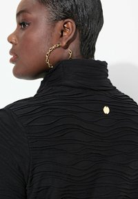 Schwarzes Oberteil mit strukturiertem wellenförmigen Muster, hohem Kragen und goldenen Hardware-Details. Große goldene Kreolen runden den Look ab.
