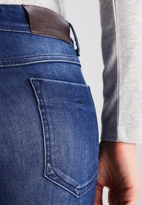Mörkblå jeans i denim har en triangulär bakficka och en brun läderlogotyp på midjan. Materialet har en strukturerad yta.