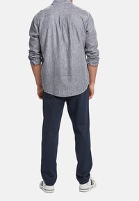 Gestreifte Button-Up-Hemd mit langen Ärmeln, blau-weißer Stoff, abgerundetem Saum, kombiniert mit dunkelblauen Hosen und weißen Sneakers.