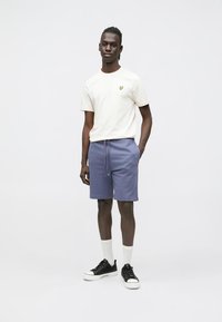 Lichtbeige T-shirt met een klein geel logo, gecombineerd met donkerblauwe shorts en zwarte sneakers. Model staat met één hand in de zak.