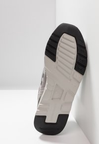 Grå mockasinsneakersula med svarta gummidetaljer, texturerade mönster och vit mellansula med upphöjda sektioner och varumärkeslogotyp.