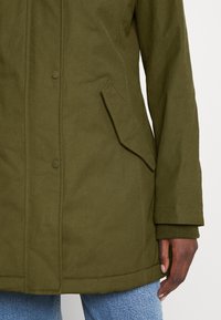 Olivengrøn polstret jakke med trykknaplukning, skrå flaplomme, manchetter på ærmerne, båret over lyseblå jeans.