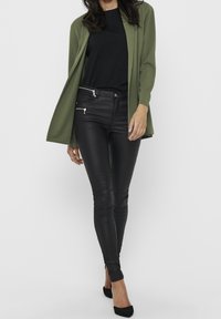 Cárdigan largo verde oliva sobre una camiseta negra, combinado con pantalones ajustados de imitación cuero negros con bolsillos con cremallera y tacones altos negros.