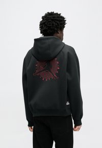 Μαύρο φούτερ με μεγάλο κόκκινο λογότυπο Jumpman στην πλάτη, με στρογγυλό γραφικό σχέδιο. Μαλακό ύφασμα και άνετη εφαρμογή με κουκούλα.