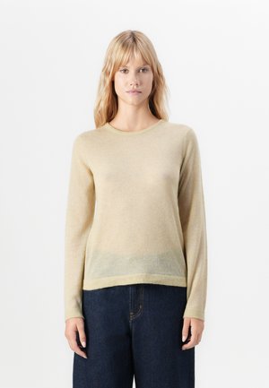 Woman standing against white background wearing a beige long-sleeve sweater and dark blue jeans, facing forward with arms relaxed.