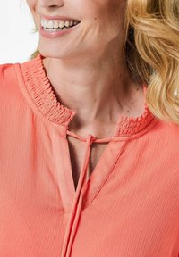 Korallenbluse aus strukturiertem Stoff mit einem gerafften Kragen und einem Schlüsselspalt-Design, das mit einem Bindeband gesichert ist.