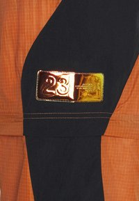 Oranžovo-černá bunda s lesklým zlatým odznakem s číslem "23". Texturovaná tkanina s kostkovaným vzorem a kontrastním šitím.