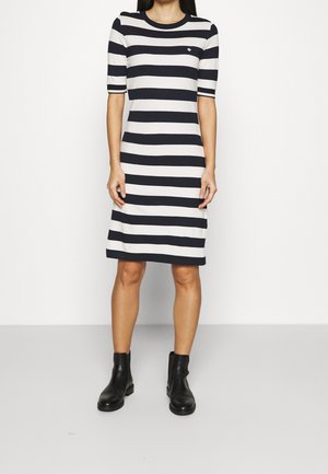 Femme portant une robe à rayures horizontales noires et blanches avec des manches mi-longues et des bottines noires, se tenant devant un fond uni.