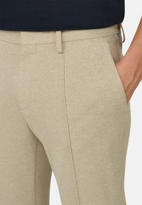 Gros plan sur un pantalon beige sur mesure avec une poche latérale, des passants de ceinture et une main partiellement dans la poche, porté avec un haut foncé.