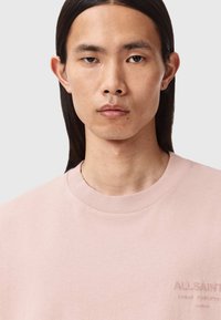 Jasnoróżowa bawełniana koszulka z okrągłym dekoltem, z wyraźnie haftowanym logo na klatce piersiowej i gładką, jednolitą fakturą.