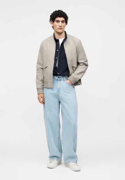 Jeune homme aux cheveux bouclés portant une veste beige, une chemise noire, un jean large bleu clair et des baskets blanches, debout avec une main dans la poche.