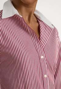 Κοντινό πλάνο ατόμου που φοράει πουκάμισο με κόκκινες και λευκές κάθετες ρίγες και λευκό γιακά, εμφανίζοντας τον λαιμό και το άνω μέρος του στήθους.