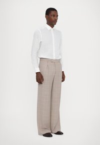 Camicia bianca in lino con bottoni abbinata a pantaloni a gamba larga grigio chiaro a quadri con accenti rosa. Il modello si trova su uno sfondo neutro.