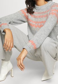 Szary sweter z dzianiny z koralowo-złotym geometrycznym wzorem. W połączeniu z jasnoszarymi spodniami i białymi botkami za kostkę. Luźny krój, ściągacze na rękawach.