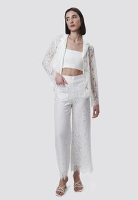 Kanten blazer met decoratieve knopen, gecombineerd met een kanten crop top en wijde kanten broek, allemaal in het wit, met franjeranden.