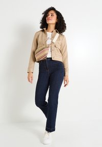 Veste beige boutonnée avec deux poches avant sur une chemise blanche, assortie à un jean bleu foncé et un sac crossbody multicolore. Baskets blanches.