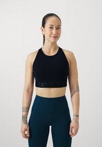 Juodas sportinis trumpas viršus su plačia juoda juosta, ant kurios užrašyta "SWEAT BETTY", derinamas su aukštu juosmeniu turkio spalvos leggingais. Lygus, elastingas audinys.