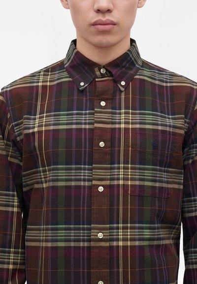 Homme portant une chemise à carreaux marron, verte et violette, boutonnée avec une poche avant et un col boutonné, visible du cou à la taille.