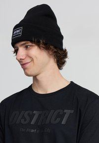 Giovane con capelli ricci che indossa un berretto nero e una maglietta nera con la scritta "DISTRICT The Heart of the City", guarda di lato con un sorriso.