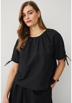 Vrouw met lang golvend haar die een losse zwarte blouse draagt met korte pofmouwen die aan de uiteinden zijn gestrikt en een kleine knoop bij de sleutelgat-halslijn.