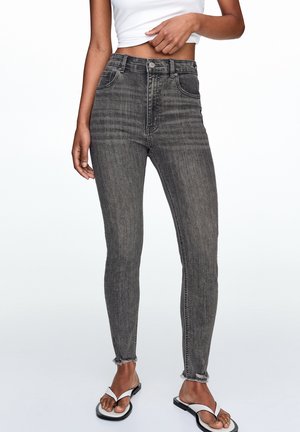 Woman wearing high-waist faded black skinny jeans with raw hem paired with a white crop top and white flip-flops on a white background.